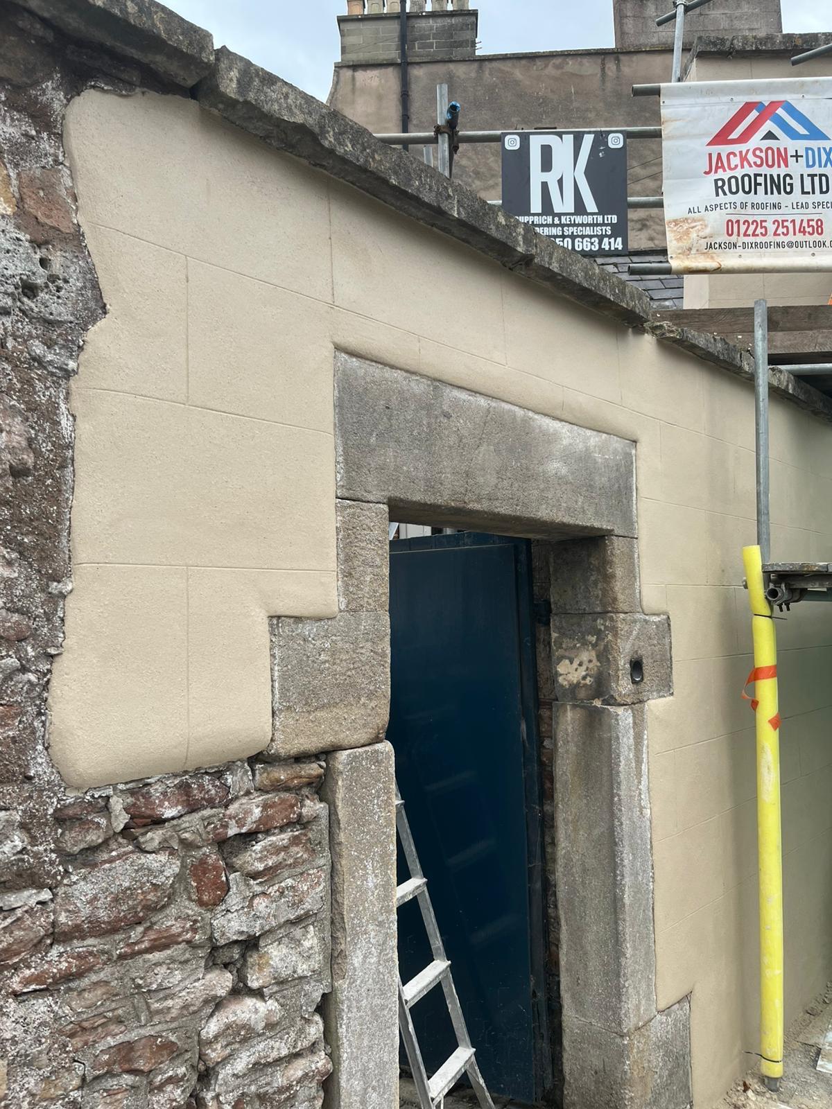 Stone doorway showing original rubble wall beside newly applied cream lime render with RK site boards