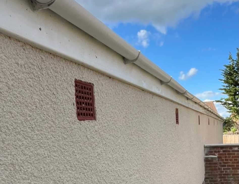 Cream roughcast rendered exterior wall with white guttering and red air bricks under blue sky